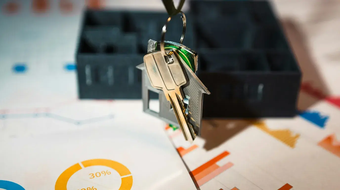 Two keys on a house-shaped keyring hanging above printed charts and a dark storage tray.
